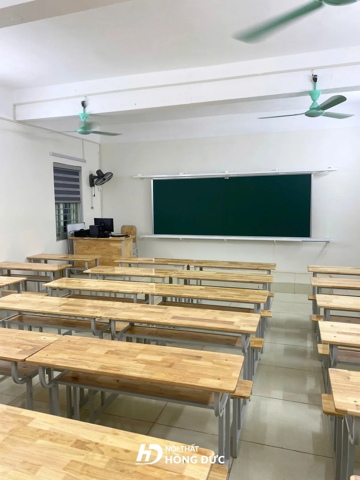 Bàn giáo viên trên bục giảng trước bảng, phía sau là các dãy bàn học sinh