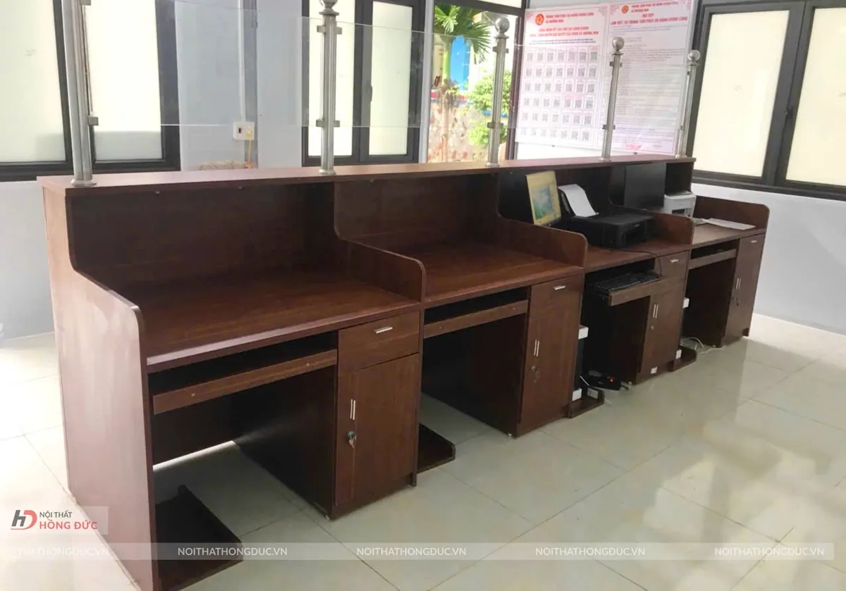 Quầy một cửa thật: mặt bàn làm việc thấp có ngăn kéo và khay bàn phím, phía sau là tấm chắn ốp gỗ và kính cao hơn hẳn